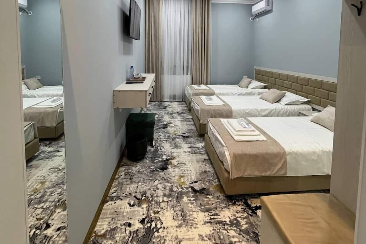 A hotel room with four beds arranged in a row, a flat-screen TV mounted on the wall, a small bedside table with a lamp, and a large window with curtains.