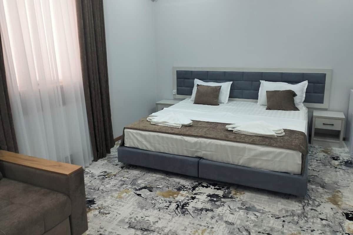 A neatly made bed with a grey headboard, white bedding, and two pillows. There is a bedside table with a lamp, a small drawer, and a window with blinds. The floor has a patterned carpet.