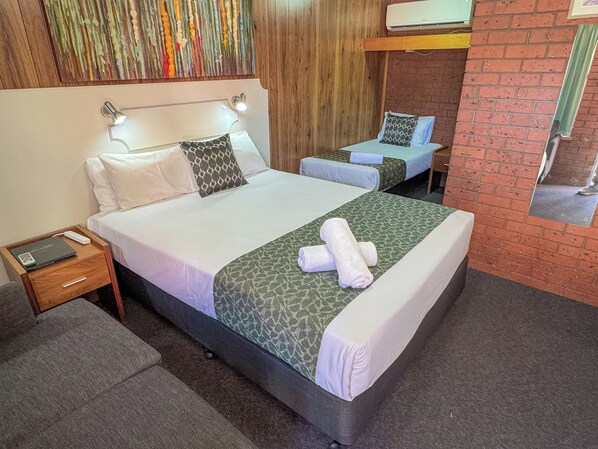 A hotel room with a large bed, two smaller beds, a wooden headboard, a colorful artwork, and a brick wall. - Pastoral Hotel Motel Echuca CBD (Echuca)