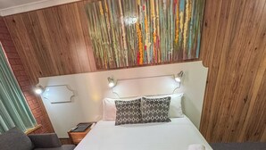 A hotel room with a large bed, two pillows, a green patterned bedspread, a wooden headboard, a nightstand with a lamp, and a grey sofa.