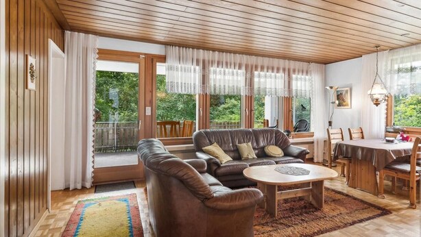 A spacious living room with a wooden ceiling, large windows with sheer curtains, a brown leather sofa, a round coffee table, and a dining area with wooden chairs and a table. - Vacation home with parking lot Pet-friendly in the ski area (Wildhaus-Alt Sankt Johann)