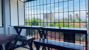 A covered outdoor seating area with tables and benches, a view of greenery and buildings through a metal fence. - Emerald Coast Hotel Negombo (Kochchikade)