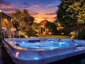 Un jacuzzi con chorros de agua, rodeado por una terraza de madera, sillas y un jardín con árboles y un edificio de ladrillo al fondo.