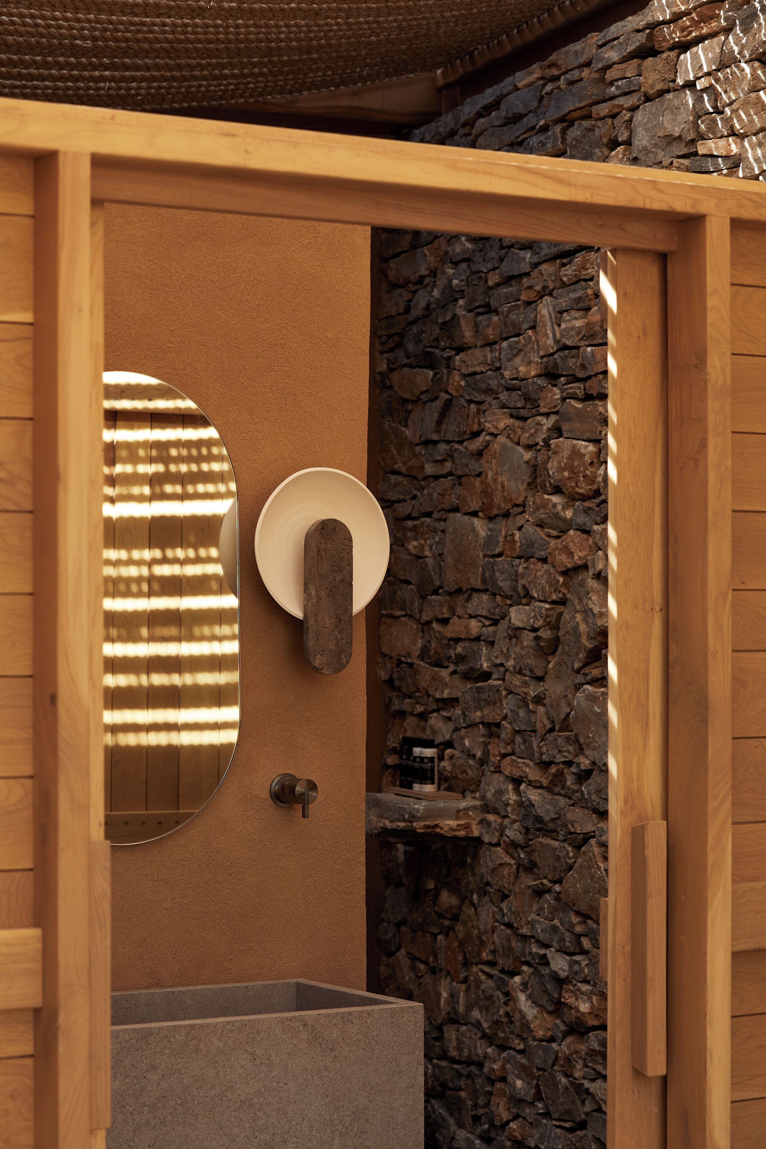 Un baño con pared de piedra, puerta de madera, espejo y lavabo.