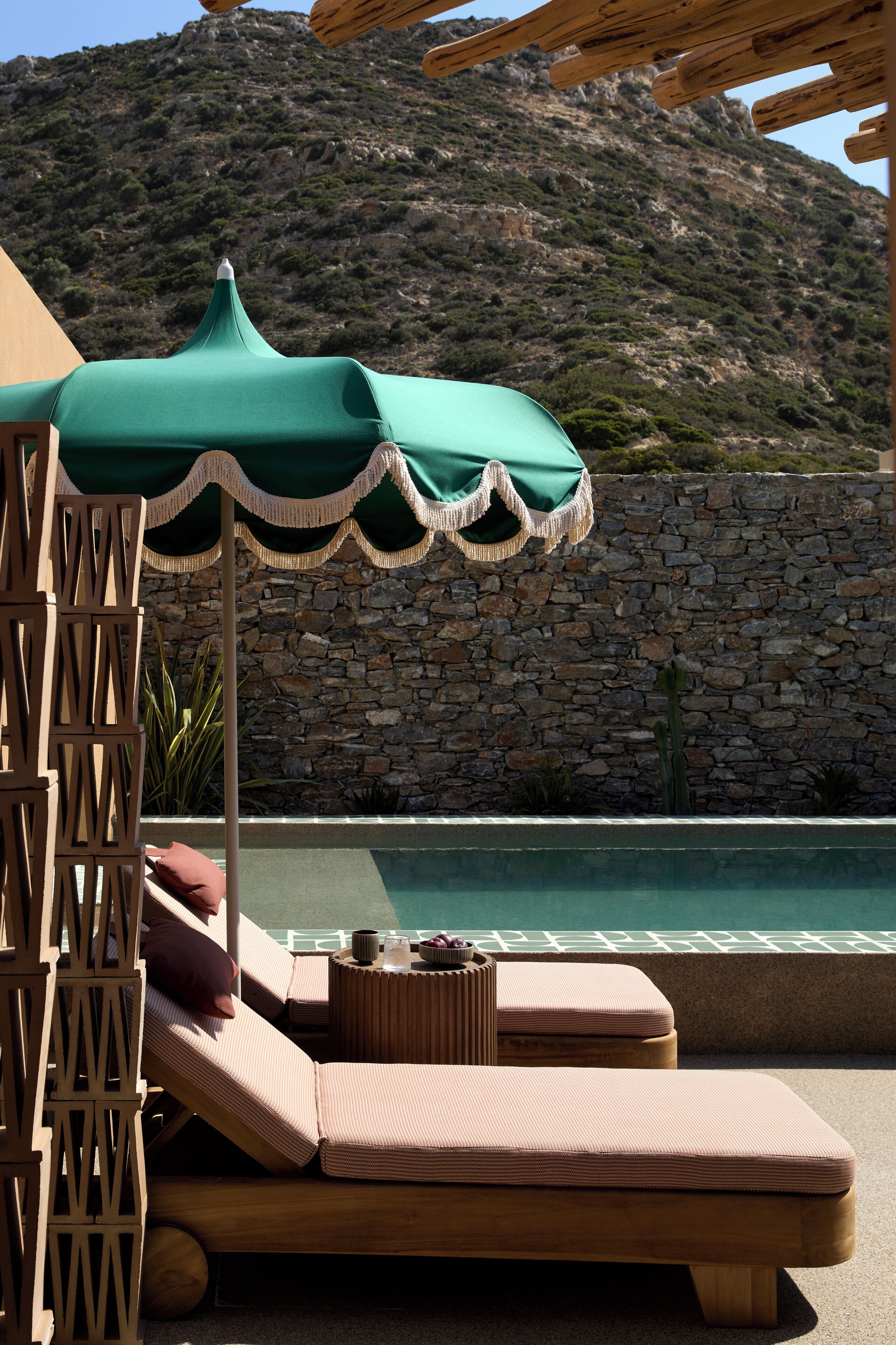 Un área de descanso junto a la piscina, con un muro de piedra, una valla de madera y una sombrilla verde.