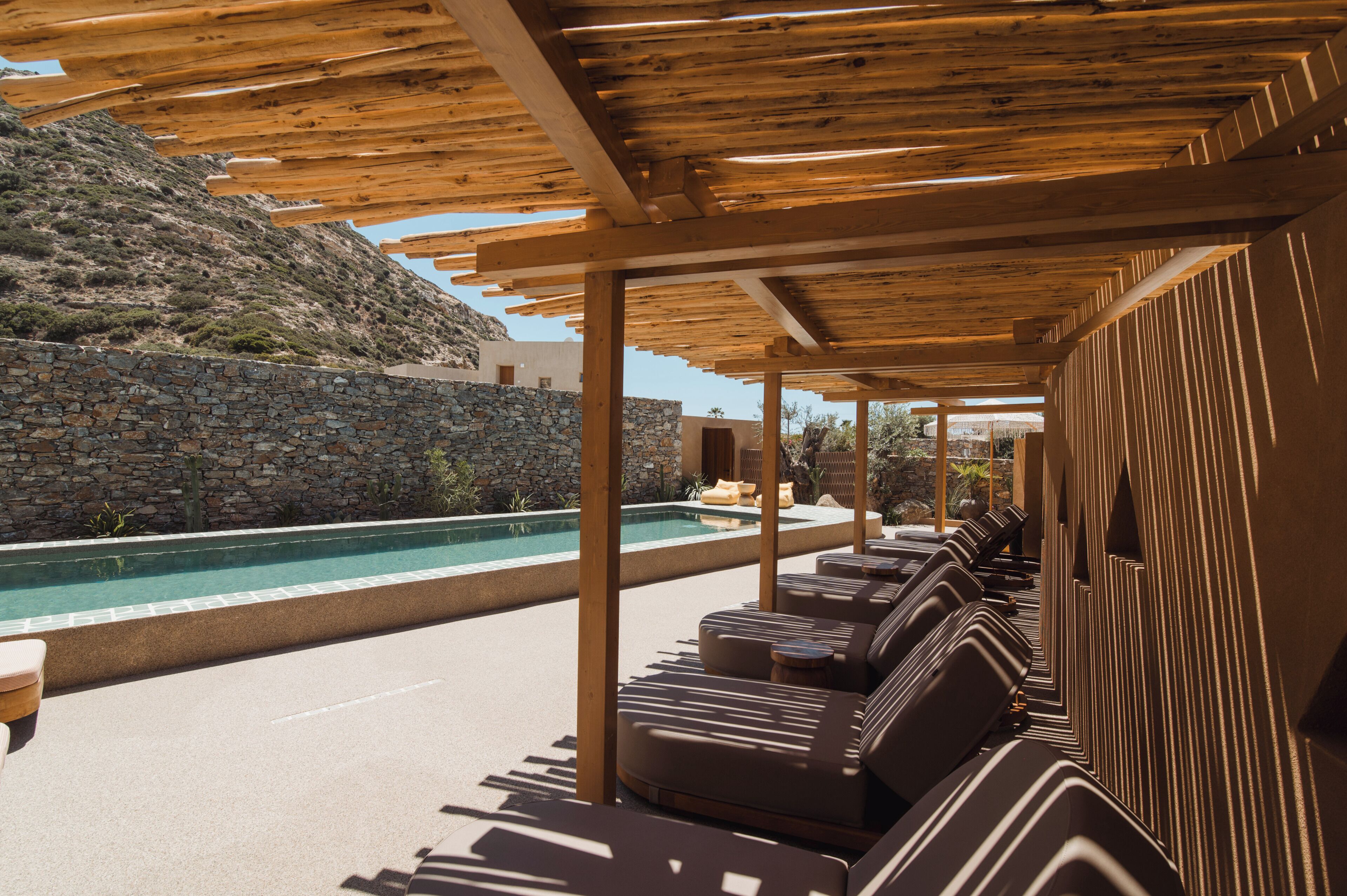 A poolside area with wooden pergolas, lounge chairs, and a stone wall.