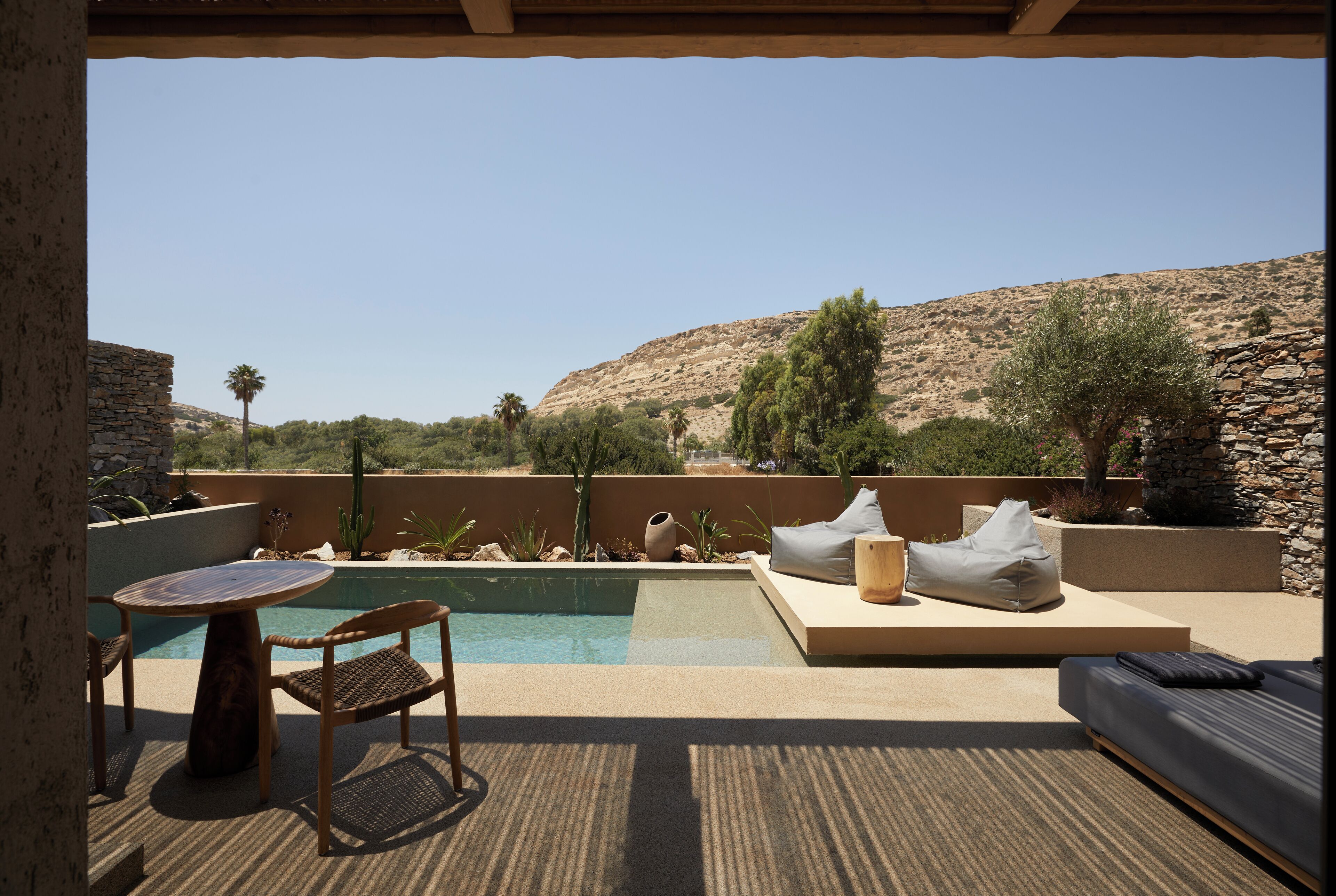 Un área junto a la piscina con un muro de piedra, una mesa de madera, una silla de mimbre y un espacio de descanso con cojín y un taburete de madera.