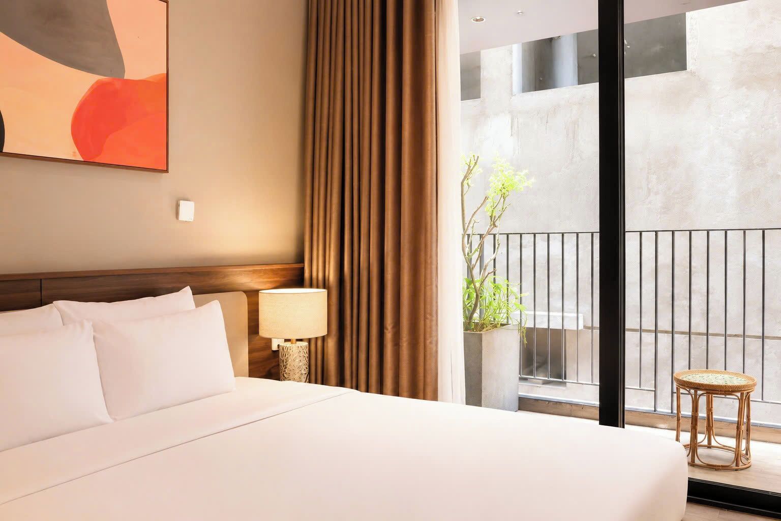 A hotel room with a large bed, a bedside table with a lamp, a view of the balcony, and a painting on the wall.