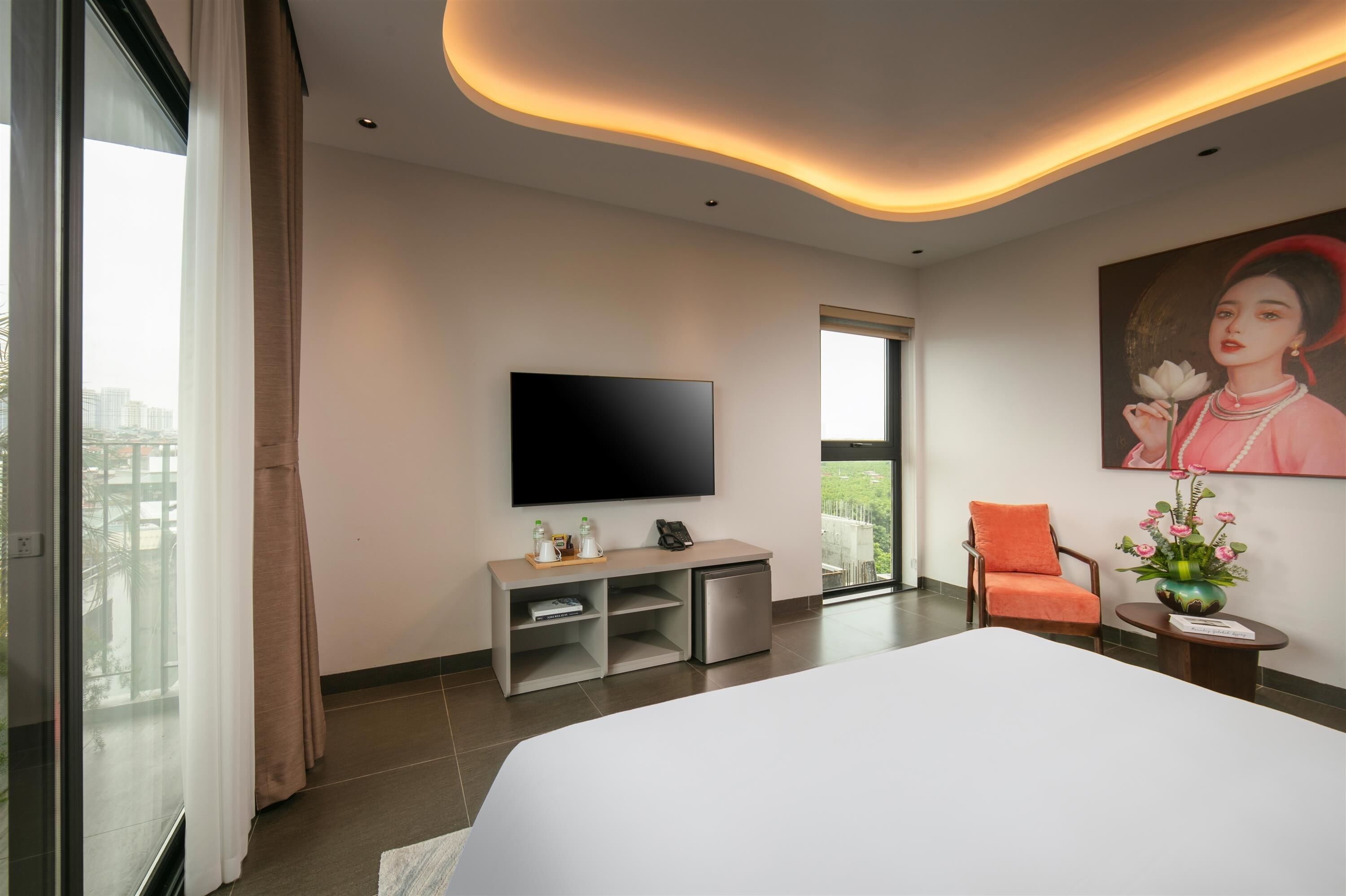 A modern hotel room with a large bed, a flat-screen TV, a small desk with a phone and a vase of flowers, a chair, and a large window offering a city view.