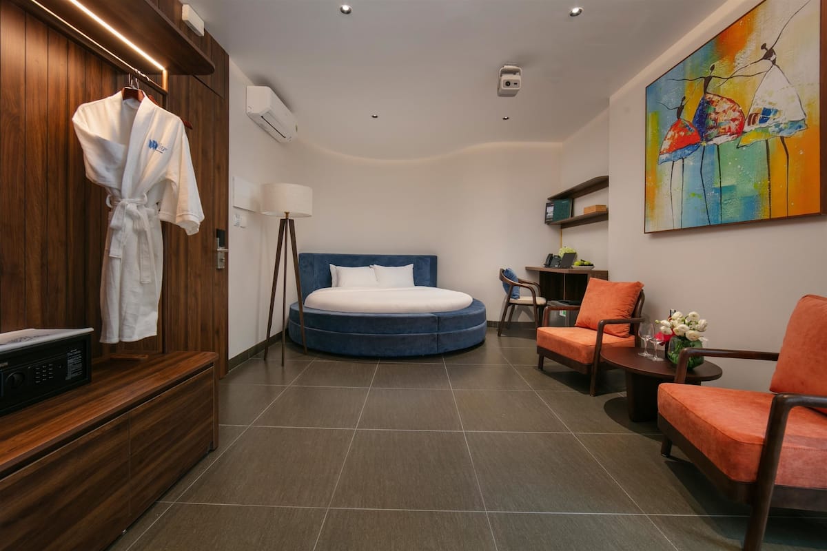 Una habitación de hotel moderna con una cama redonda, una zona de estar con sillas naranjas, un armario de madera y un cuadro en la pared.