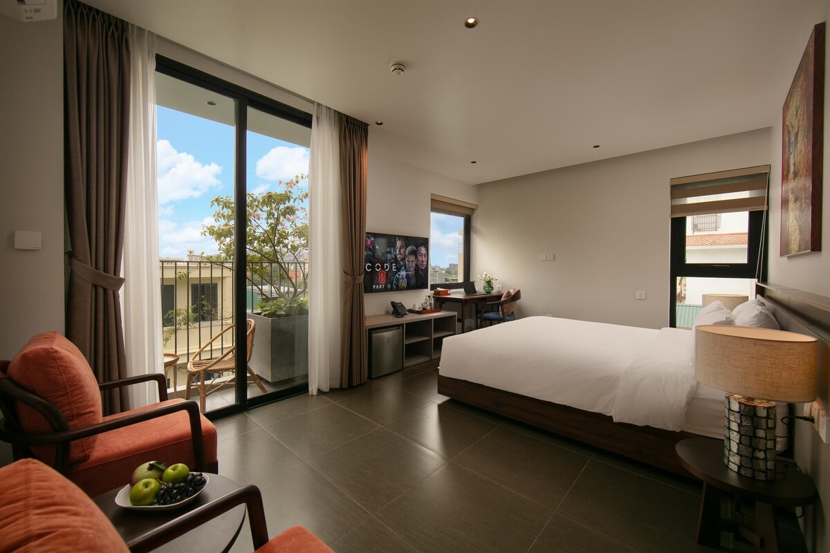 A modern hotel room with a large bed, a desk with a laptop, a TV, and a balcony with curtains.