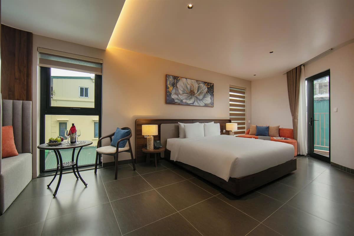 Una habitación de hotel con una cama grande, una mesita con frutero, una silla, una mesita de noche con lámpara y un cuadro en la pared.