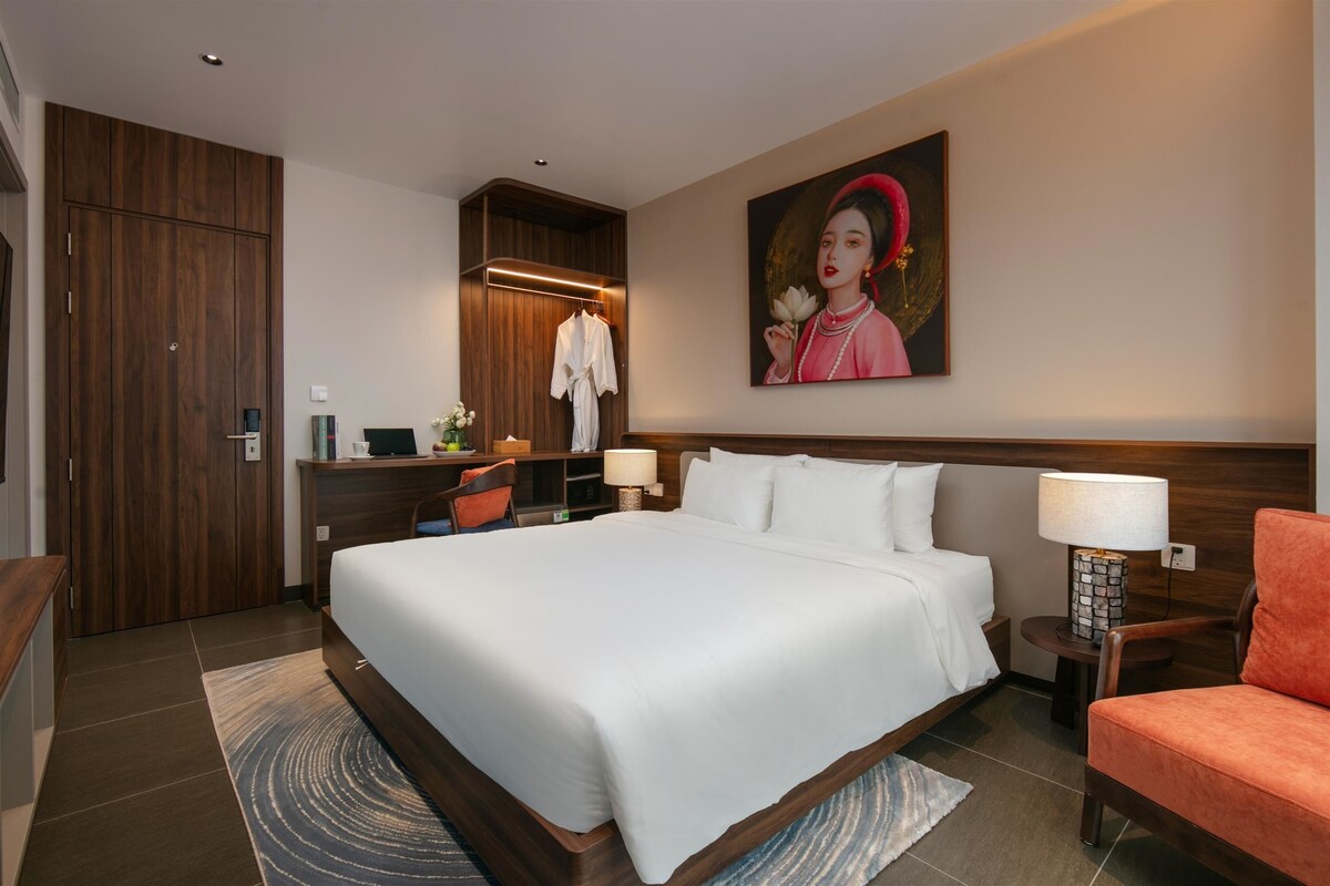 A hotel room with a large bed, a wooden wardrobe, a desk with a chair, a nightstand with a lamp, and a painting on the wall.