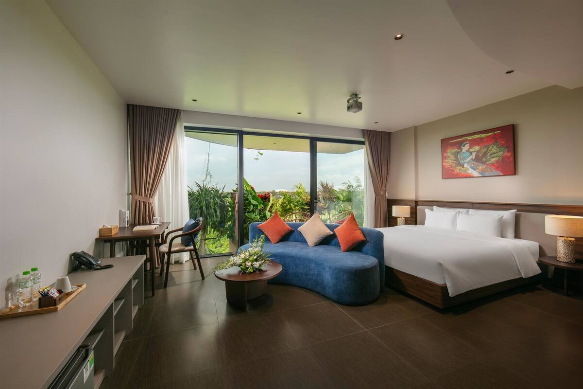 Habitación de hotel moderna con sofá azul curvo, cama grande, escritorio con silla y vistas a zonas verdes a través de amplios ventanales.
