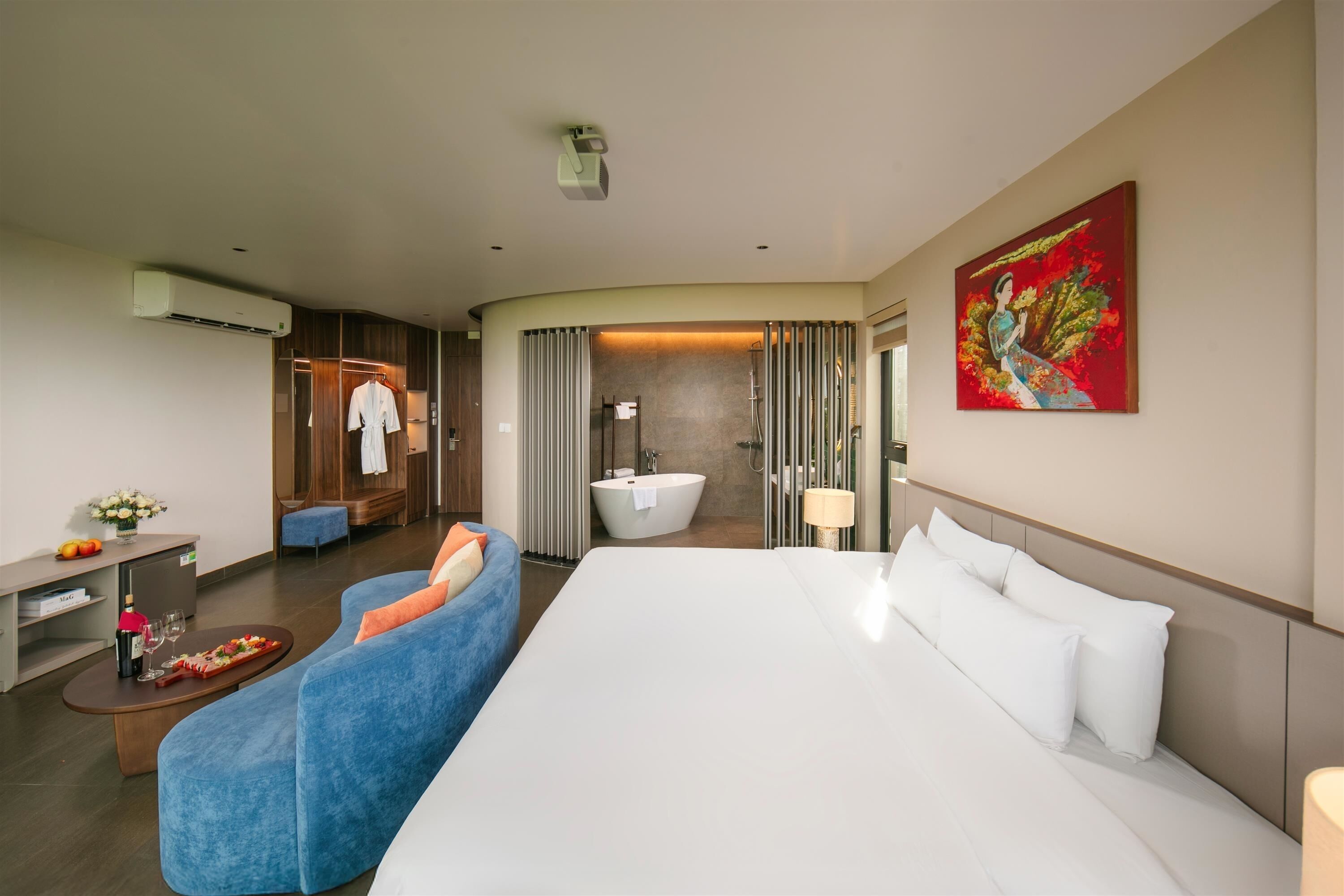 Una camera d'albergo moderna con un letto grande, un angolo salotto con un divano blu, un tavolino con una bottiglia e dei bicchieri, un accappatoio appeso, un quadro al muro e un bagno con vasca e doccia.