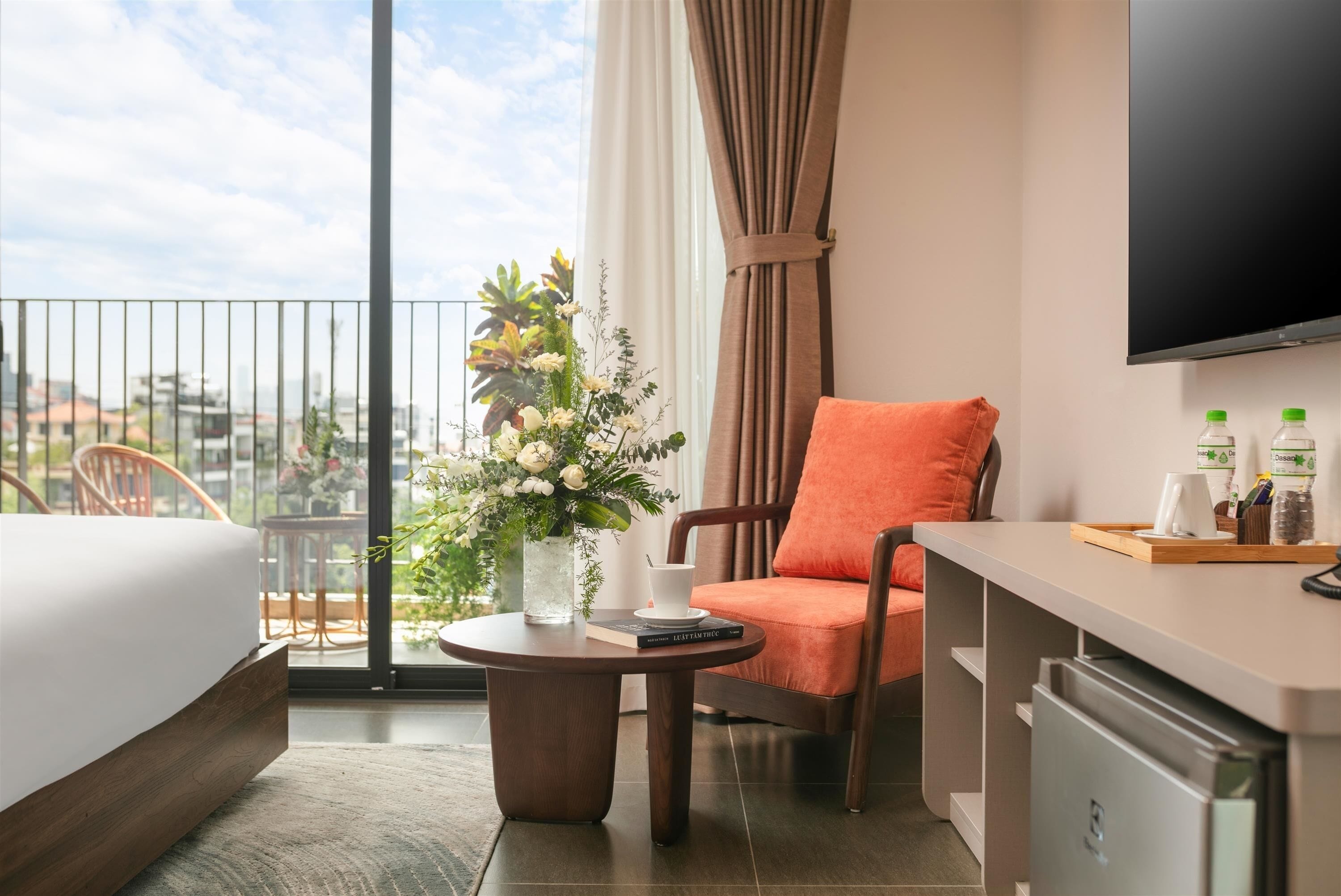 A hotel room with a bed, a chair, a small table with a vase of flowers, a television, and a minibar.