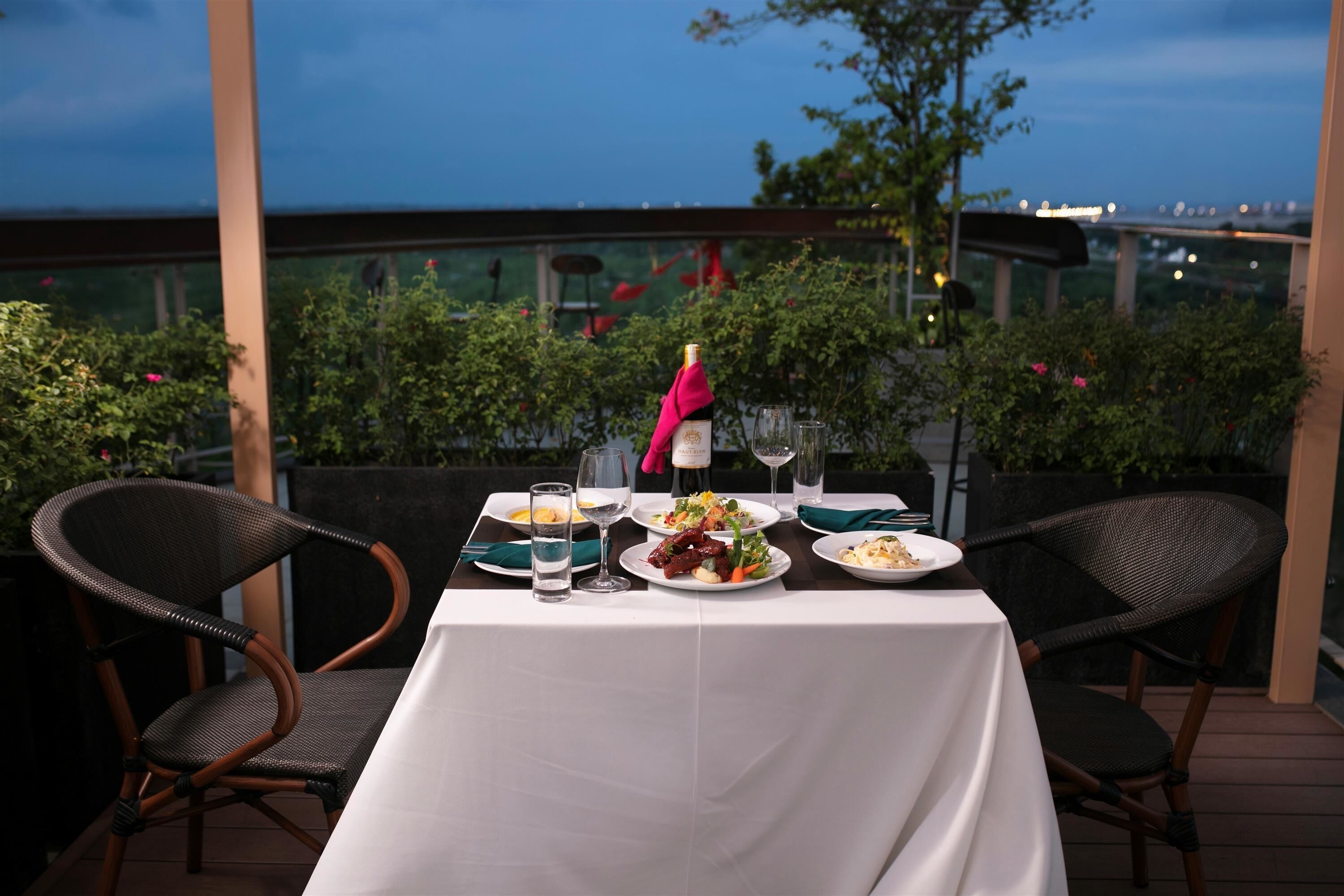 An outdoor dining table set with plates, glasses, and a bottle of wine, surrounded by wicker chairs.