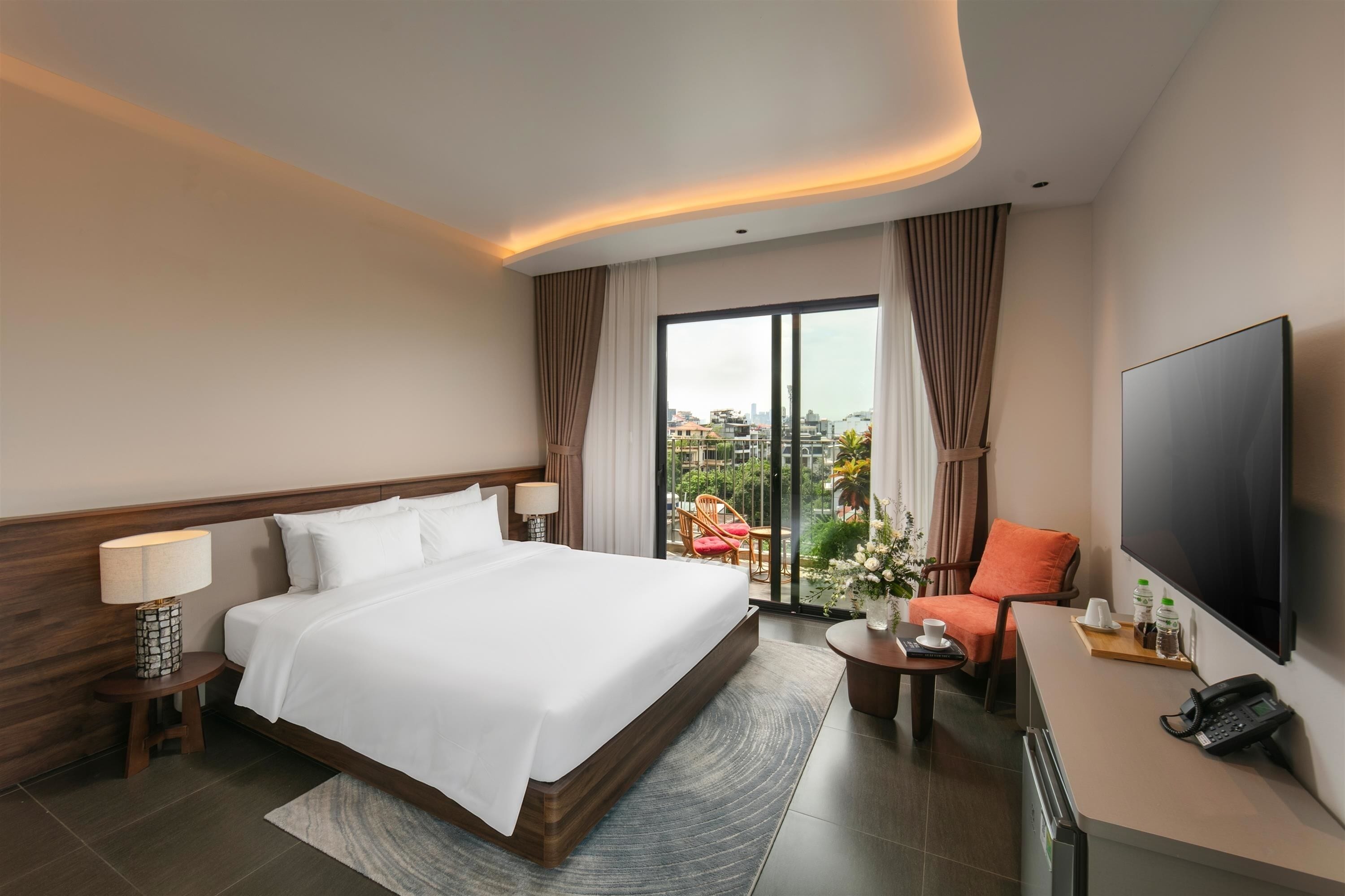 A hotel room with a large bed, a desk with a telephone, a TV, and a view of the city through the window.