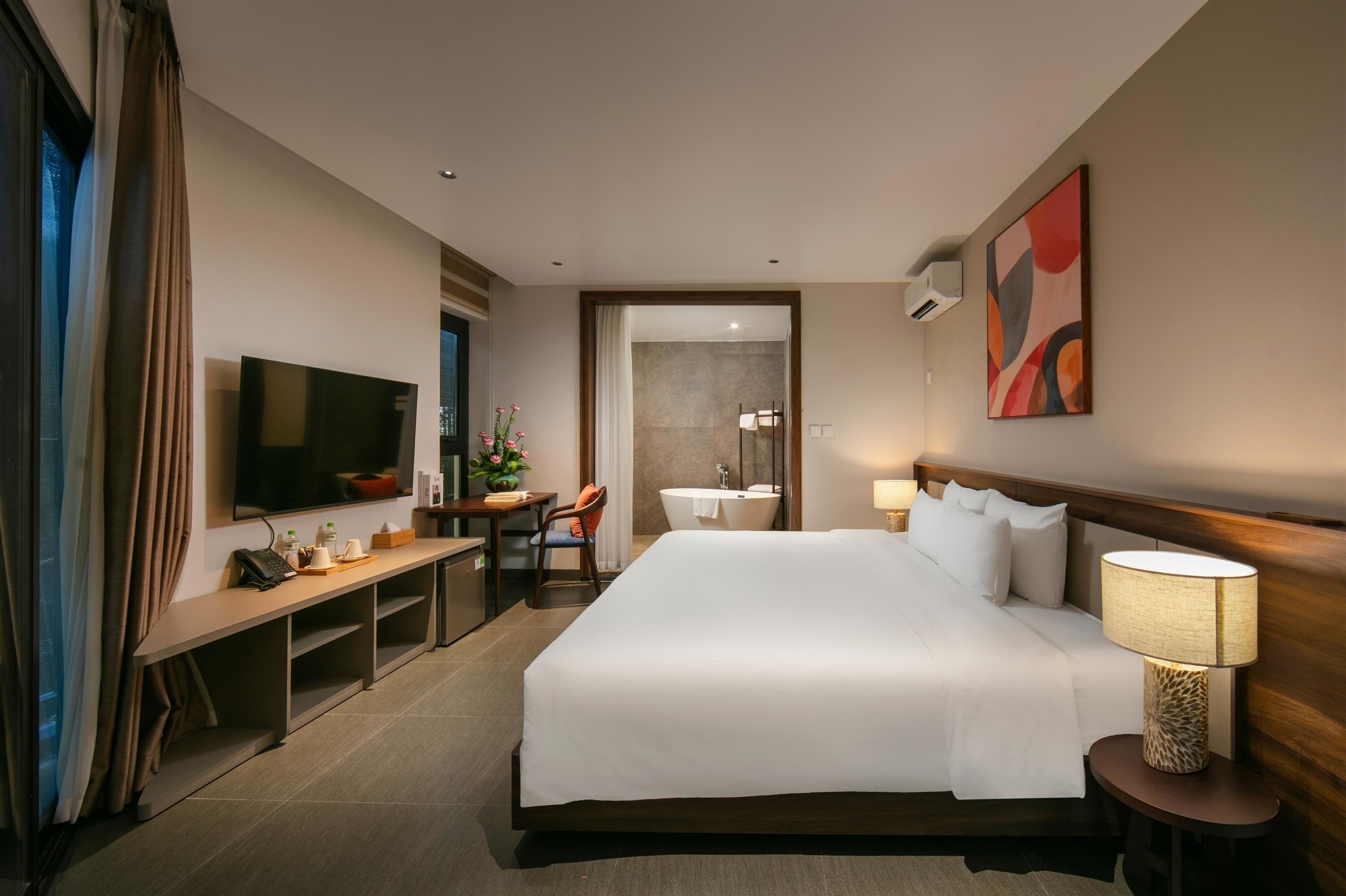 A hotel room with a large bed, a desk with a chair, a television, a bathroom with a sink and mirror, and a painting on the wall.