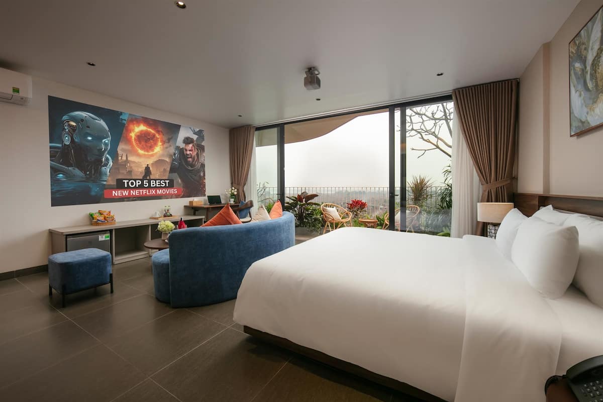 Habitación de hotel moderna con una cama grande, un televisor mostrando 'TOP 5 BEST NEW NETFLIX MOVIES', un balcón con plantas y vistas a la ciudad.