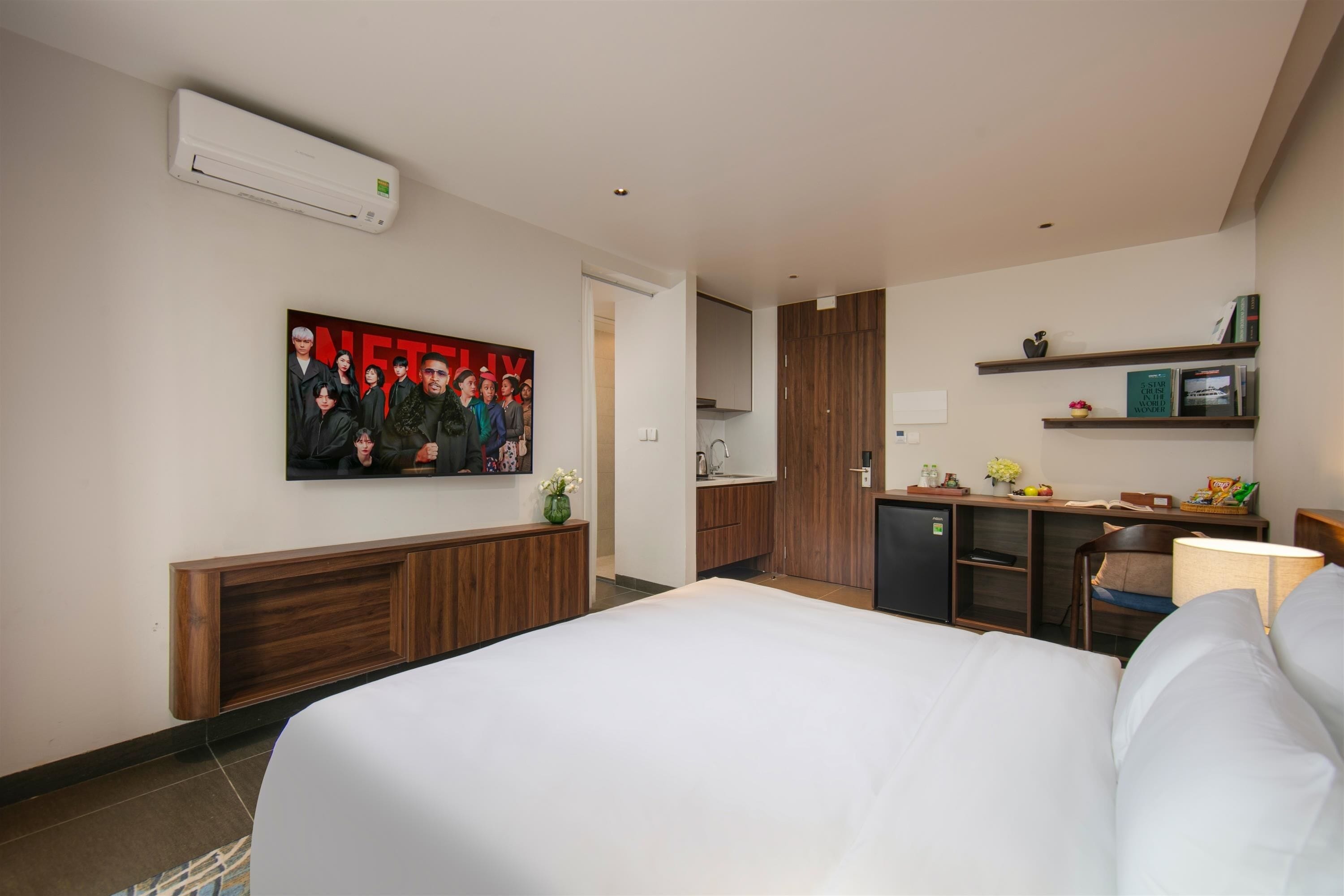 A hotel room with a large bed, a kitchenette, a wooden cabinet, a wall-mounted air conditioner, and a framed picture on the wall.