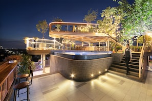 Un área de bar moderna al aire libre con jacuzzi, asientos y una escalera que lleva a una terraza superior.
