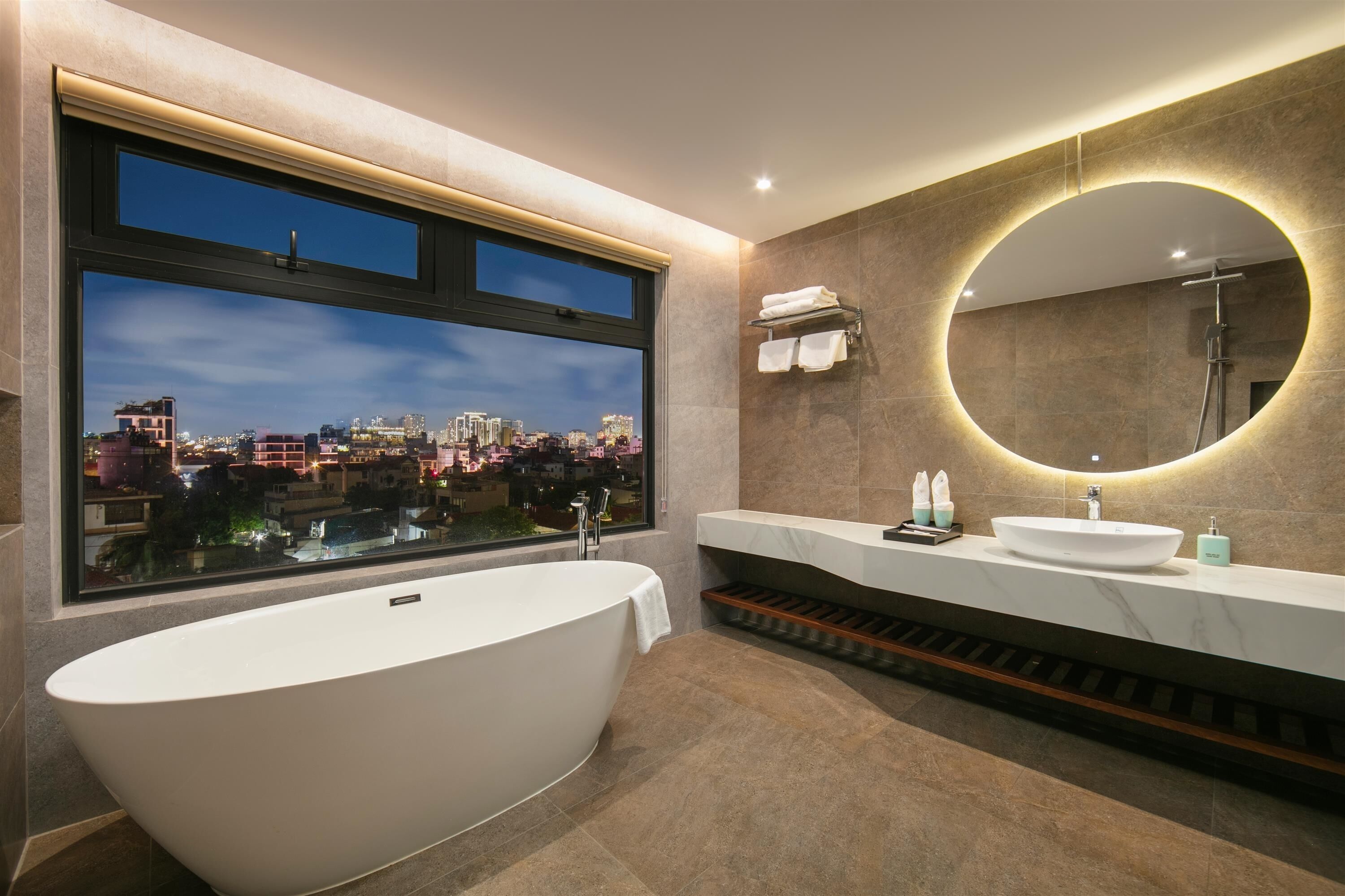 Une salle de bain moderne avec une baignoire sur pieds, une grande fenêtre donnant sur la ville, un grand miroir rond avec éclairage intégré et un meuble-lavabo blanc doté d’un évier et d’un porte-serviettes.
