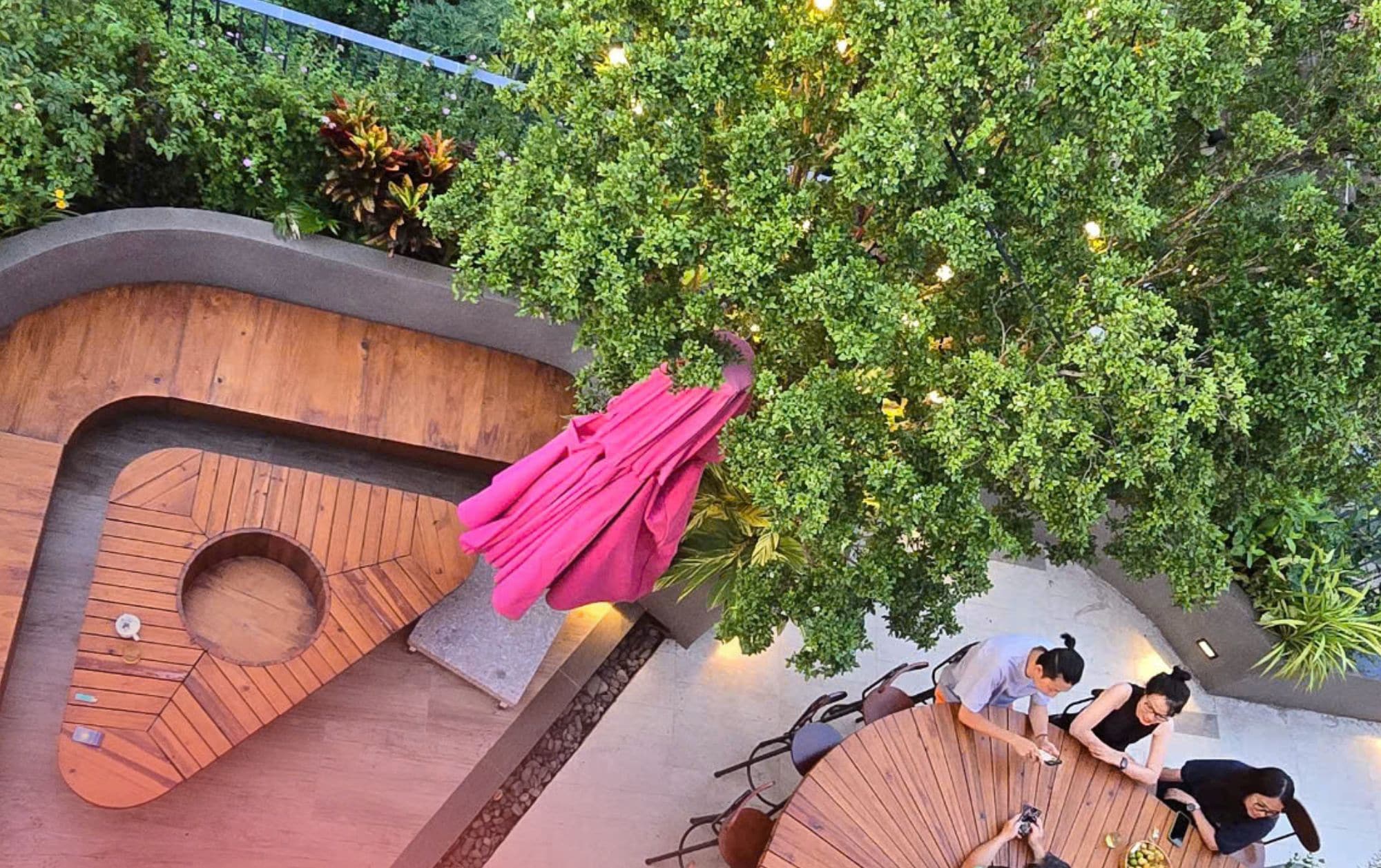 An outdoor seating area with a wooden table and chairs, surrounded by greenery and a tree.