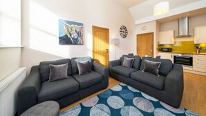 A modern living room with two grey sofas, a colorful abstract painting, a clock, and a kitchen in the background.