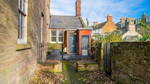 A narrow alleyway leading to a small red-brick house with a dark door, surrounded by stone walls and a metal fence.