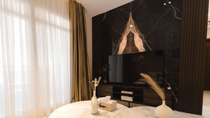 Una sala de estar moderna con piso de mármol, un televisor de pantalla plana montado en una pared oscura, una mesa redonda con un florero y una caja de pañuelos, y un ventanal con cortinas translúcidas.
