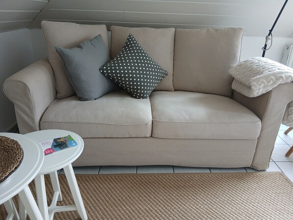 A beige sofa with two pillows, a white table with magazines, and a woven rug.