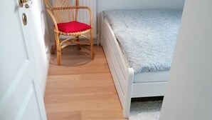 Une pièce étroite avec un plancher en bois, un lit simple recouvert d’une courtepointe à motifs, une chaise en osier avec un coussin rouge et un radiateur blanc.