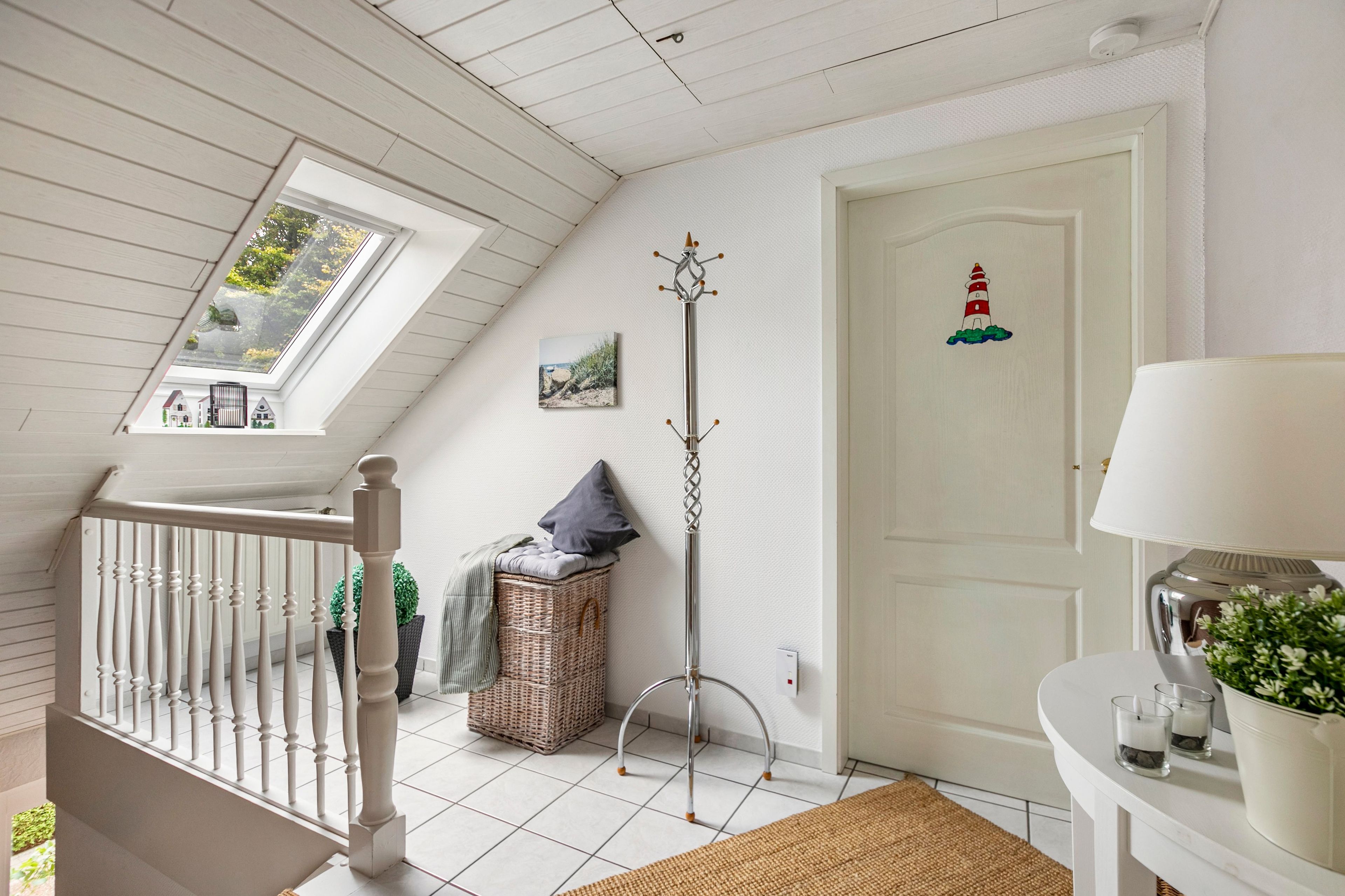 Ett rum med ett sluttande tak, en vit dörr med en bild av en fyr, en klädhängare, en korg av rotting och ett litet bord med glas och en växt.