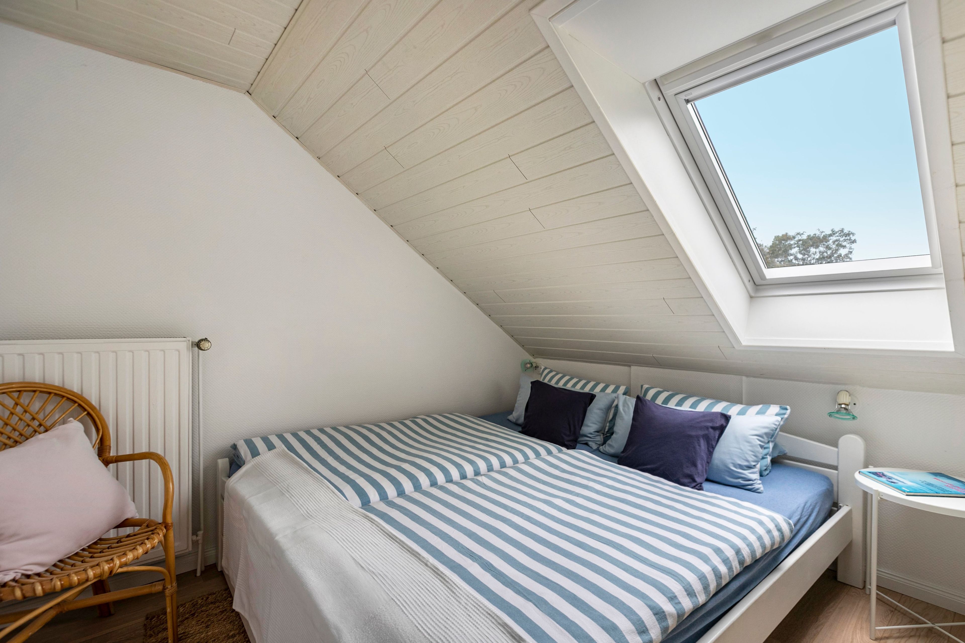 A bedroom with a sloped ceiling, a window, a bed with striped bedding, a wicker chair, and a small bedside table.