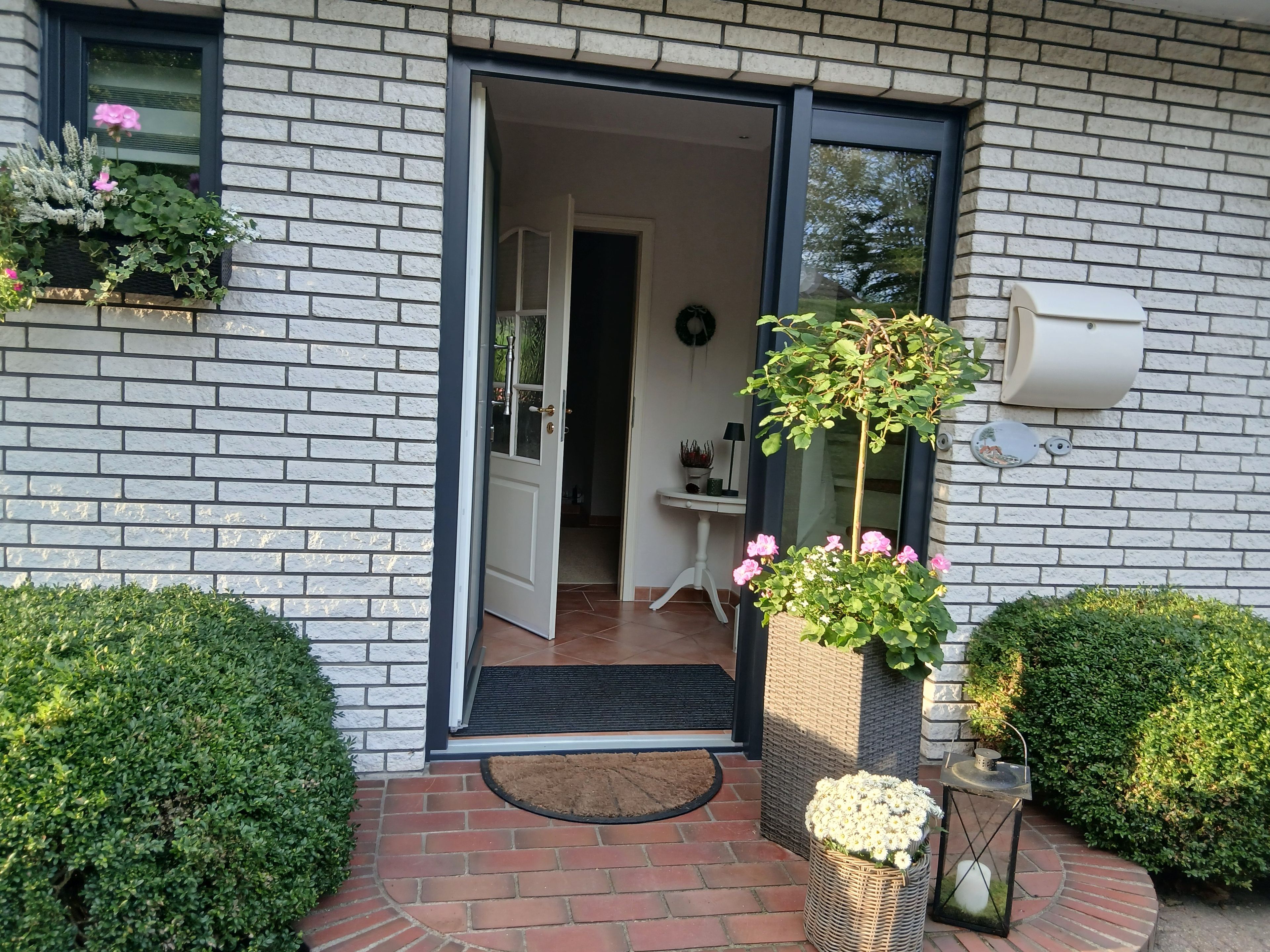 Een gevel van baksteen met een deur die uitkomt op een binnenruimte, een klein terras met een tafel en stoelen, en een bloeiende plant in een pot.