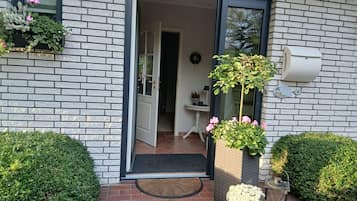 Een gevel van baksteen met een deur die uitkomt op een binnenruimte, een klein terras met een tafel en stoelen, en een bloeiende plant in een pot.