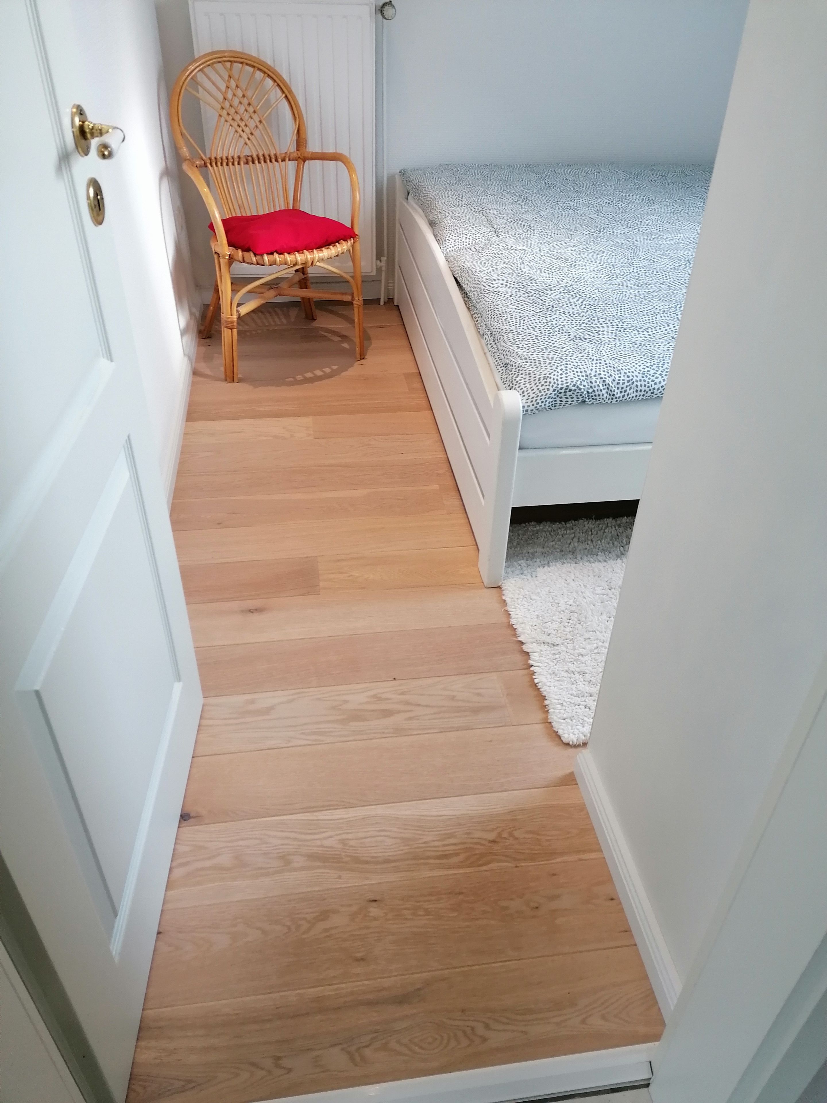 Ein schmales Zimmer mit Holzboden, einem Einzelbett mit gemustertem Überwurf, einem Korbstuhl mit rotem Kissen und einem weißen Heizkörper.