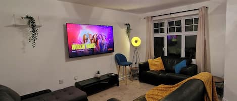 A living room with a flat-screen TV, black leather sofas, a coffee table, a side table, a floor lamp, and a window with curtains.