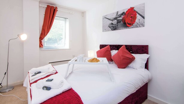 A hotel room with a neatly made bed, a bedside table with a tray of breakfast, a red throw blanket, and a painting of a rose on the wall. - Coventry 2-bedroom Apartment-pet Friendly (Coventry)