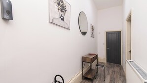 A hallway with a wooden floor, a black door, a small wooden table with a stool, and a wall-mounted hand sanitizer dispenser. - Tranquil Derby 2 Bedroom Apartment Retreat (Derby)