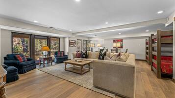 A spacious living room with a large sofa, a coffee table, and a fireplace. There are large windows with a view of trees and a balcony.
