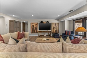 A spacious living room with a large sofa, a coffee table, and a TV. There are paintings on the wall and a lamp on the side. - Spectacular views | Private Hot Tub | Sleeps 16 (Beaver Creek)