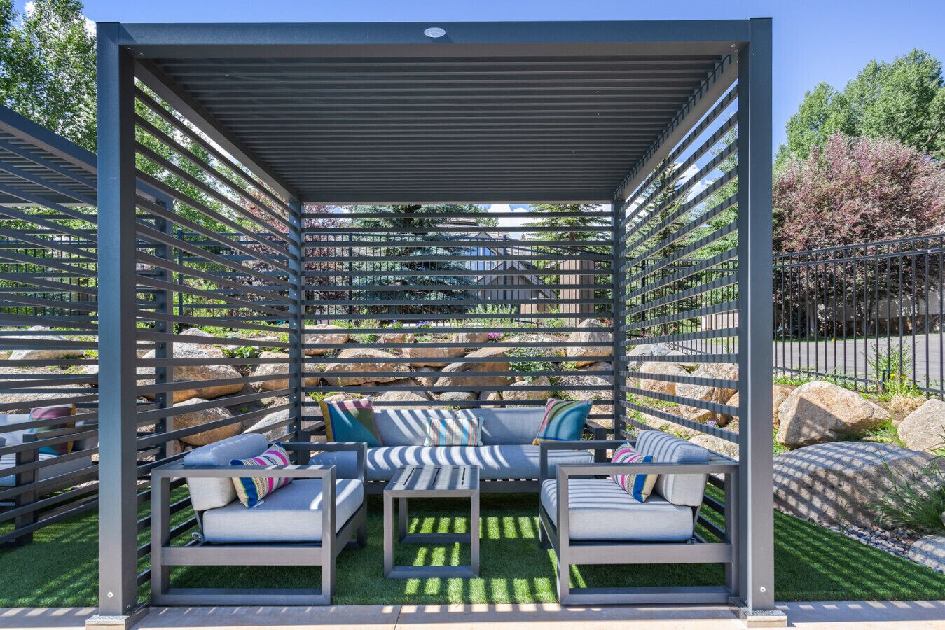 A covered outdoor seating area with multiple sofas and cushions, surrounded by a metal fence and greenery.