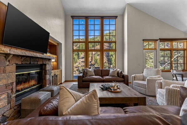A spacious living room with a stone fireplace, leather sofas, a flat-screen TV, and large windows offering a view of a forested area.