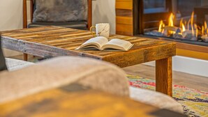 A cozy living room with a wooden coffee table holding an open book and a mug, a comfortable sofa, and a fireplace with visible flames. - New | Prime Location | Skier`s Paradise (Avon)