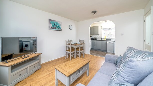 A modern living room with a grey sofa, wooden coffee table, and a TV on a wooden stand. A dining area with a wooden table and chairs is visible. The kitchen is in the background with white cabinets and a dishwasher. - Pet-friendly 1-bedroom Apartment in Crawley (Crawley)