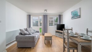 A modern living room with a grey sofa, a wooden coffee table, a TV on a wooden stand, and a dining table set with plates and glasses. - Pet-friendly 1-bedroom Apartment in Crawley (Crawley)