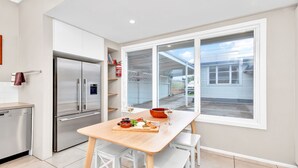 Uma cozinha moderna com geladeira de aço inoxidável, uma mesa de jantar de madeira com uma tigela de frutas e queijos, e uma grande janela com vista para uma garagem.