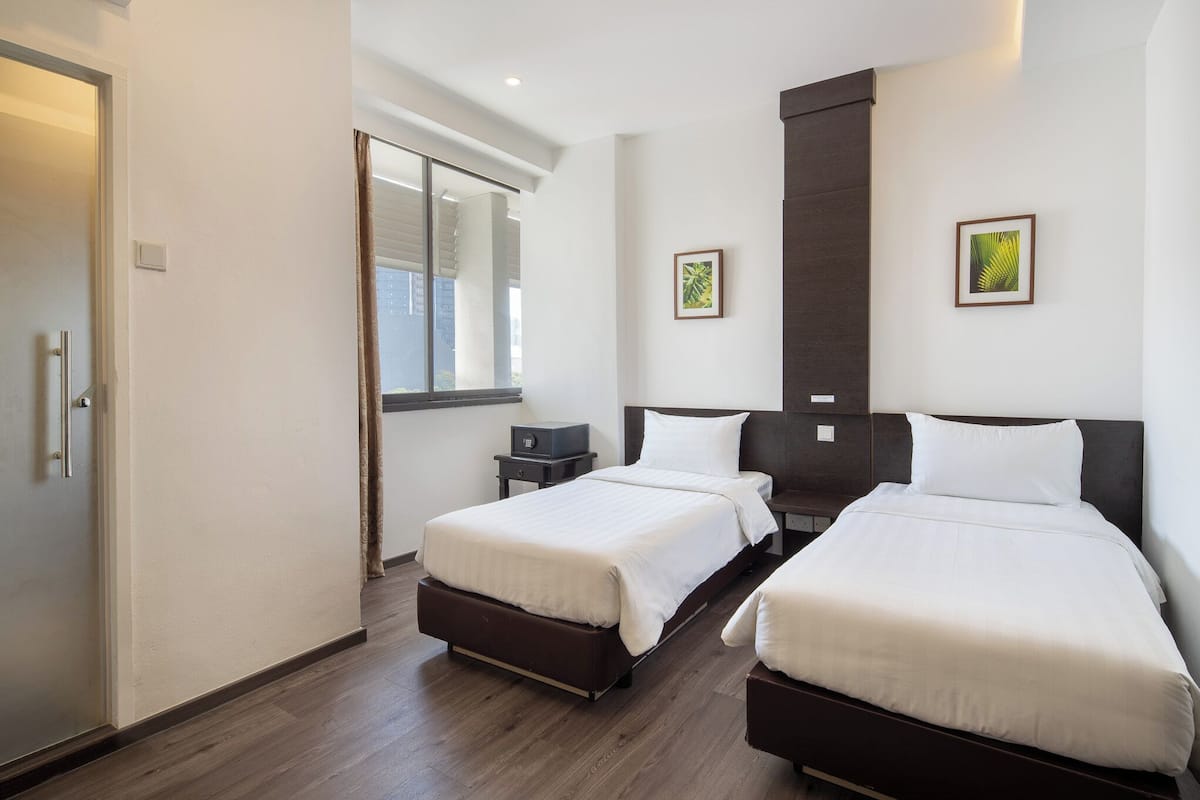 A hotel room with two beds, a window with blinds, a small bedside table, and a wall-mounted artwork.